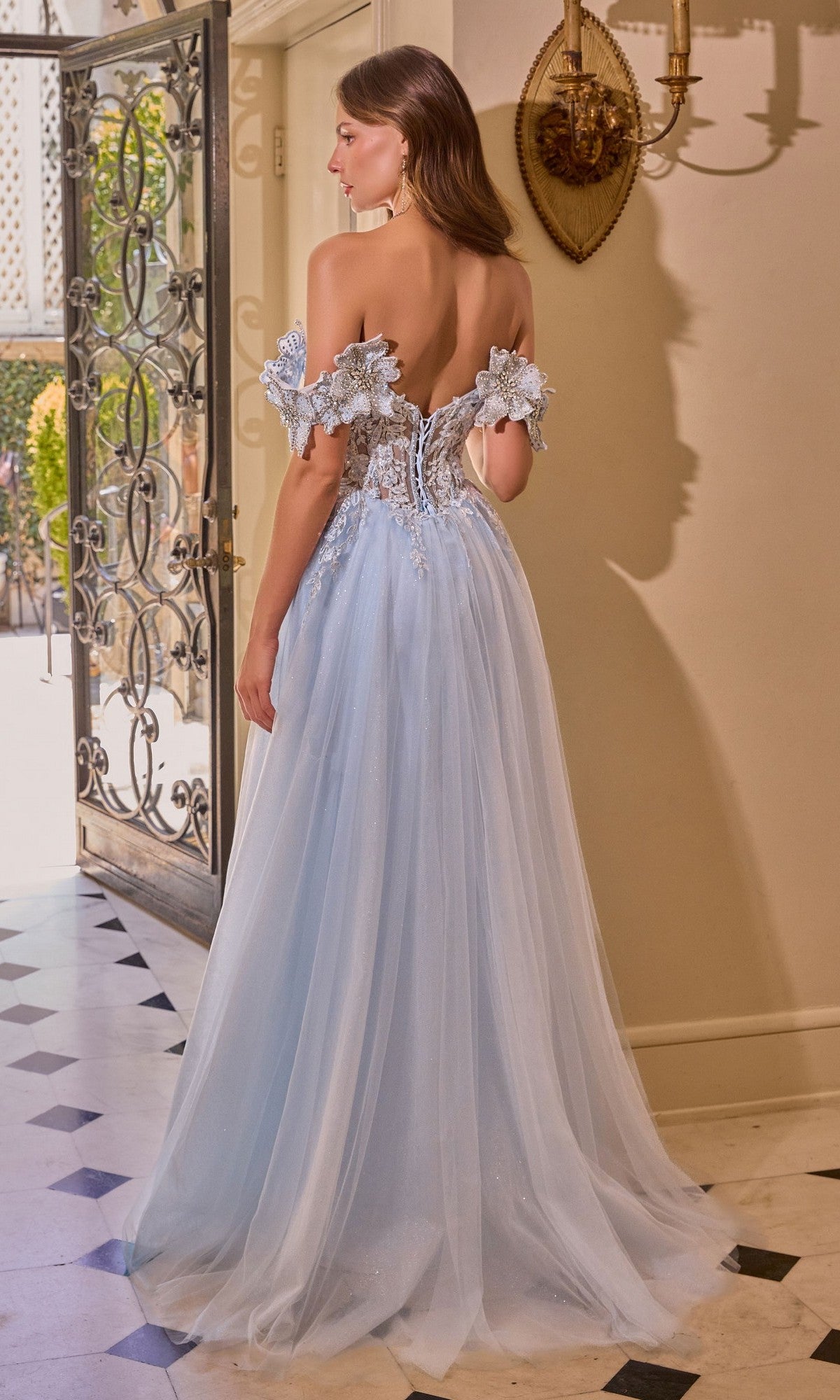 A woman in the Andrea and Leo A1512 long prom dress, featuring light blue 3D floral appliques and an off-the-shoulder a-line design, stands indoors near an open door with her back to the camera.